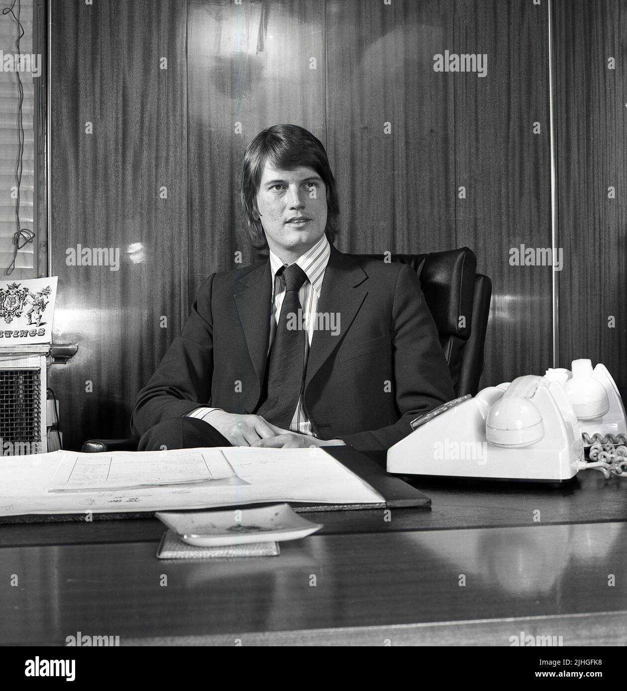 1975, historical, a young male suited car dealer sitting at his desk in his office, Croydon, South London, England, UK, with two telephones of the era. Stock Photo