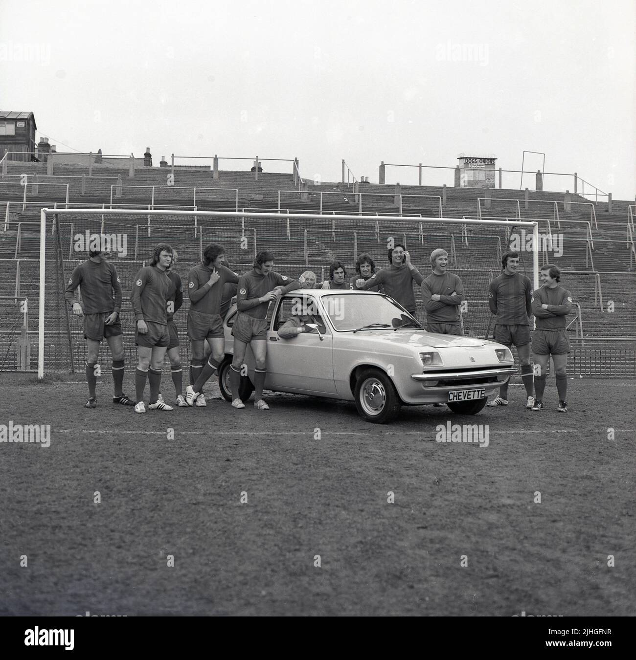 1974, historical, football manager, Malcolm Allison sitting in 2-door Chevette hatchback car on the pitch at Selhurst Park, with team players standing around. Selhurst Park is the home of Crystal Palace Football Club. A wooden hut on top of banked terraces says Police Post. A smaller on, a food stall, says Dogs & Onions. Stock Photo