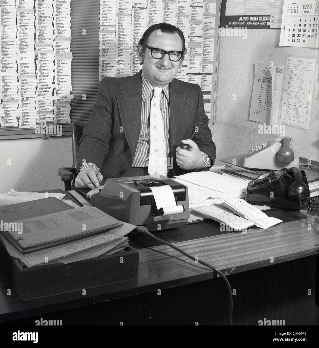 1975, historical, a manager in a car dealership sitting at his desk in his office, pipe in hand, telephones and adding machine of the era, Croydon, England, UK. A wall card index and poster for Alfred Marks Bureau, the employment agency can be seen. Stock Photo