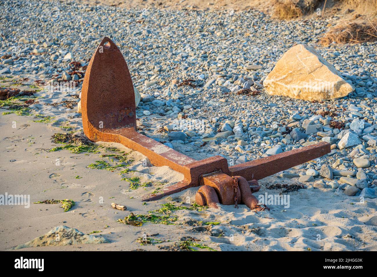 salvaged rusty shipwrecked boats anchor from the sea bed  partially buried in the sand on a rocky pebble beach Stock Photo