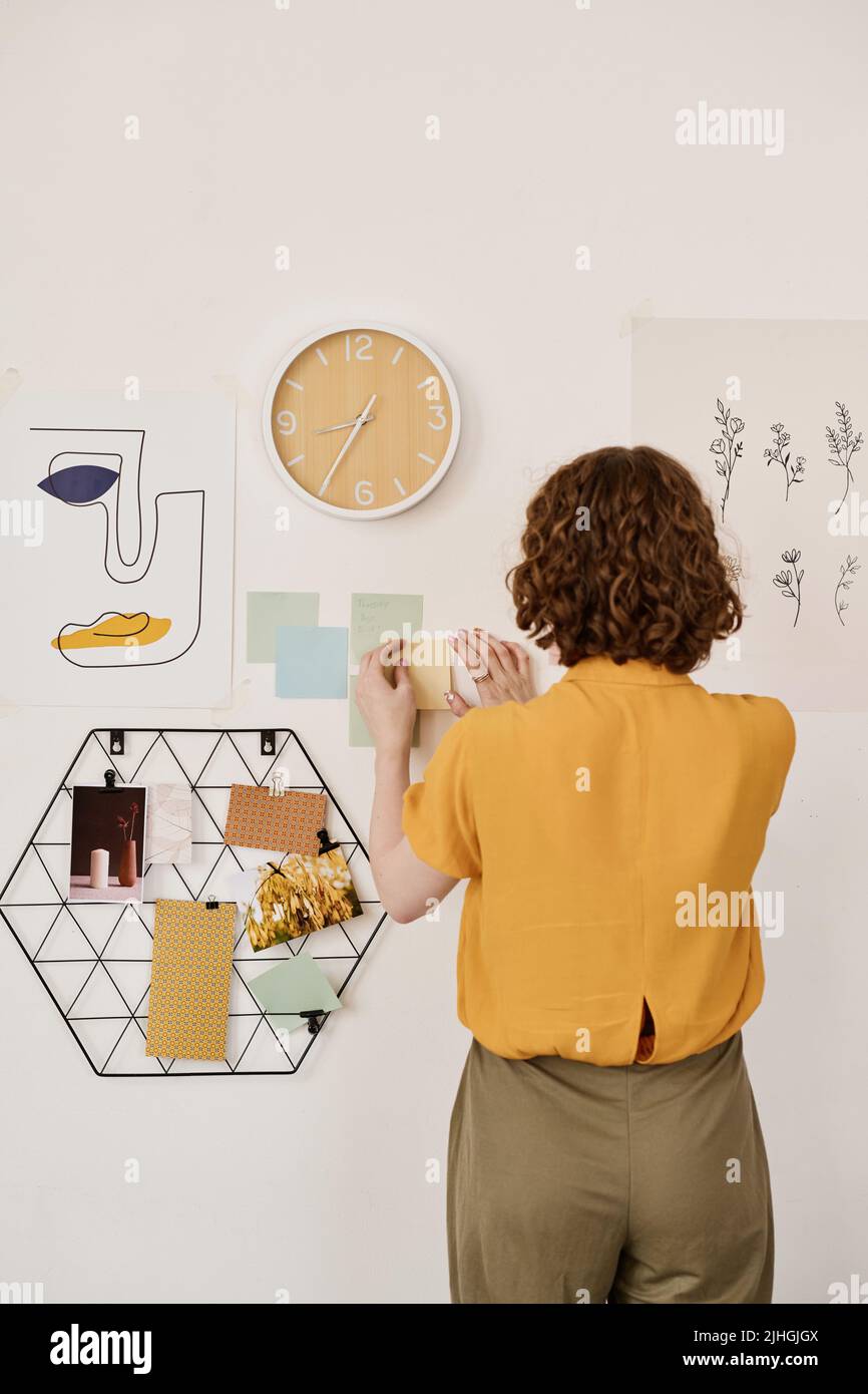 Back view of young creative female freelancer sticking notepapers with working points or plan on wall with clock, some photos and pictures Stock Photo
