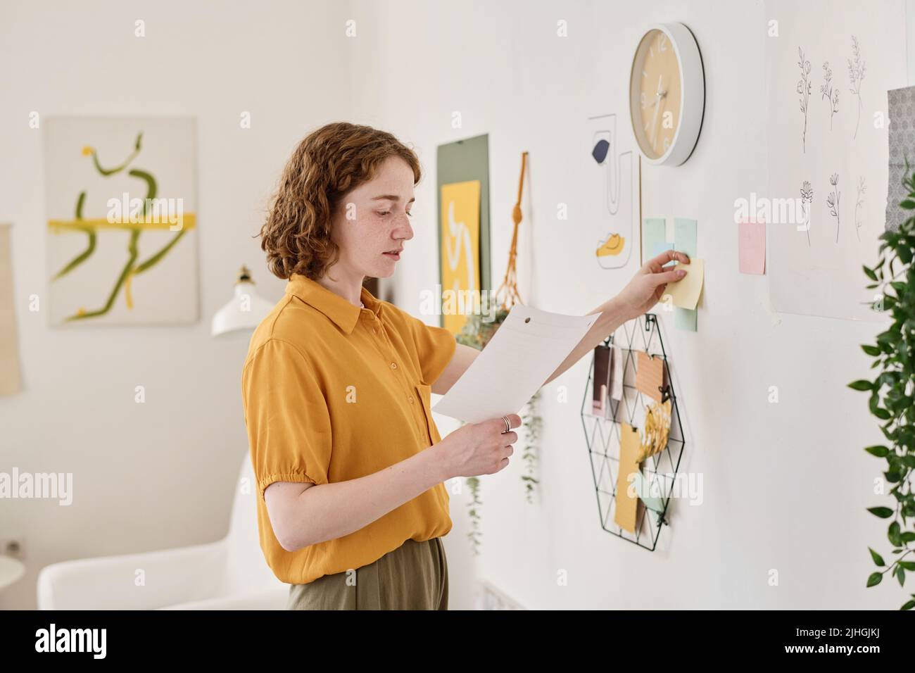 Young creative freelancer looking through information in paper document while standing in front of wall and sticking notepapers Stock Photo