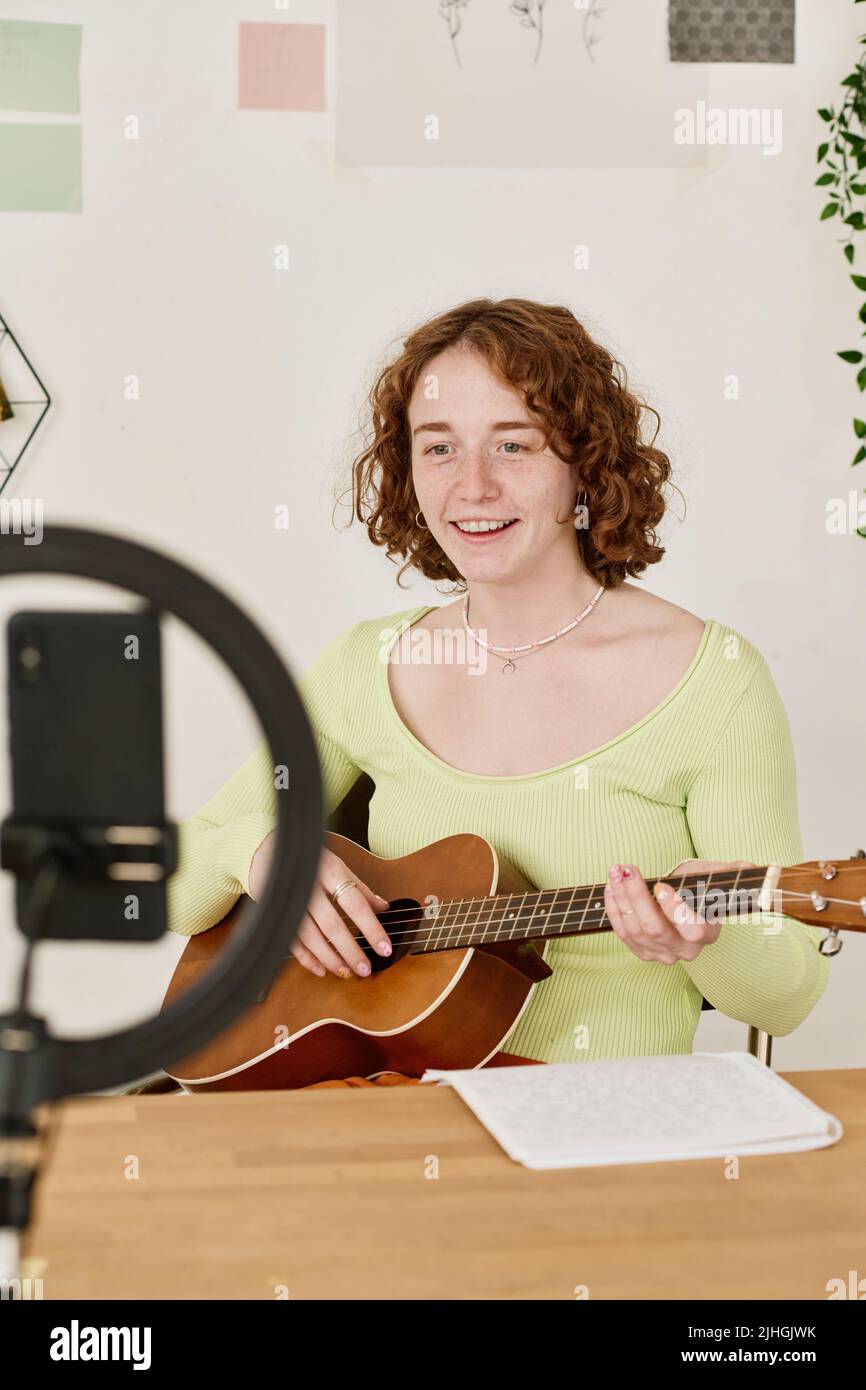 Young smiling woman with acoustic guitar making video lesson for online audience while sitting by desk in front of smartphone camera Stock Photo