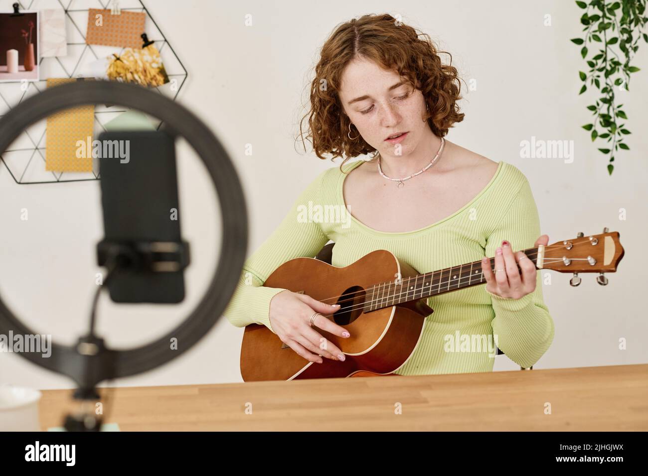 Young female student or teacher of course of guitar playing sitting by desk in front of smartphone camera during online lesson Stock Photo