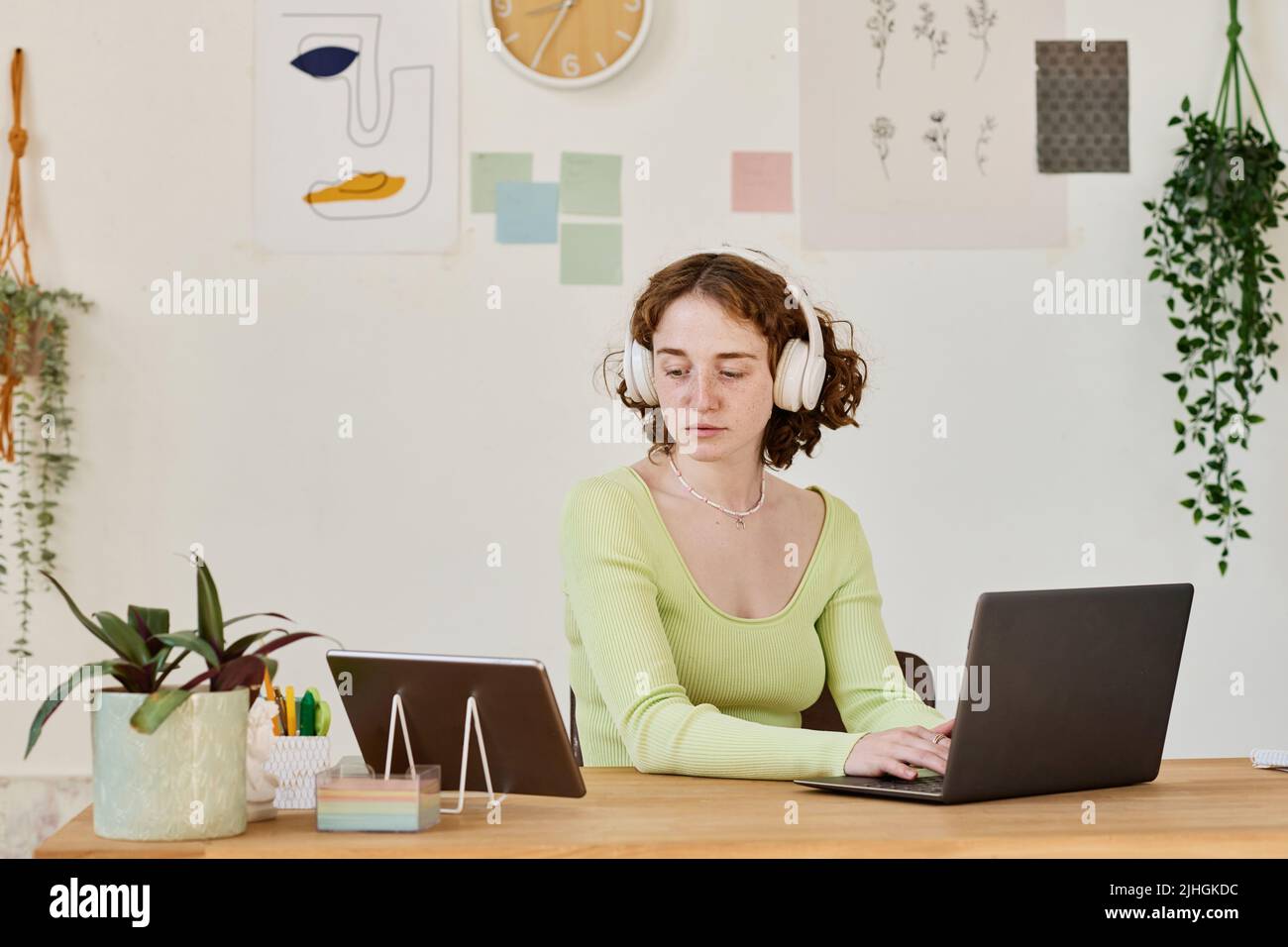 Young serious businesswoman or student in headphones networking by desk while sitting in front of laptop, typing and looking at tablet screen Stock Photo