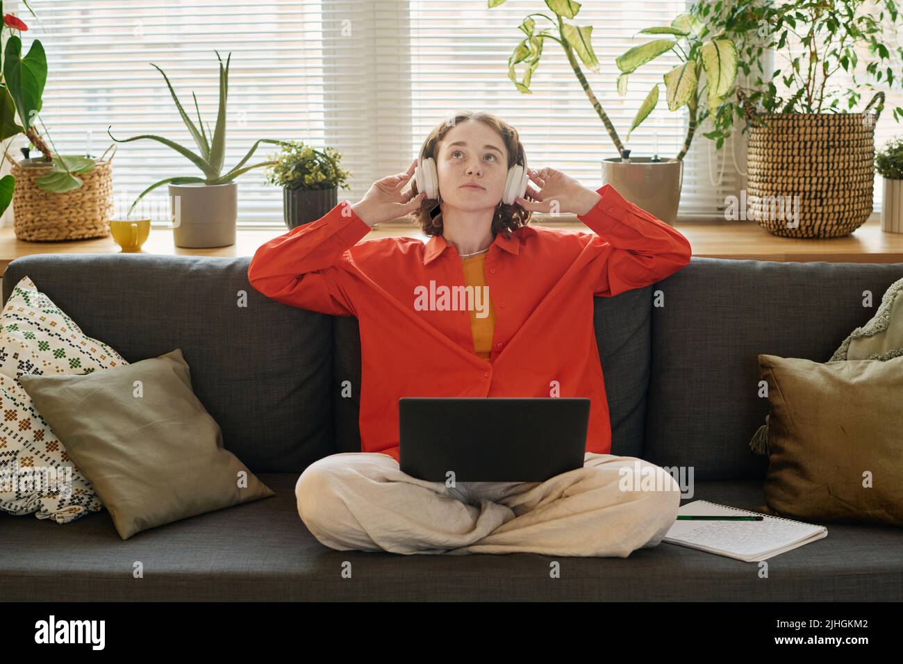 Young female student in headphones listening to audio lesson while sitting on couch in living room with laptop on her crossed legs Stock Photo