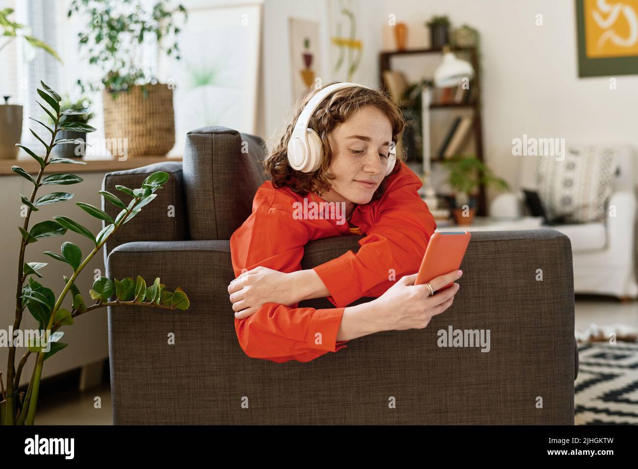Cute adolescent girl in red shirt and white headphones looking at smartphone acreen while communicating in video chat Stock Photo