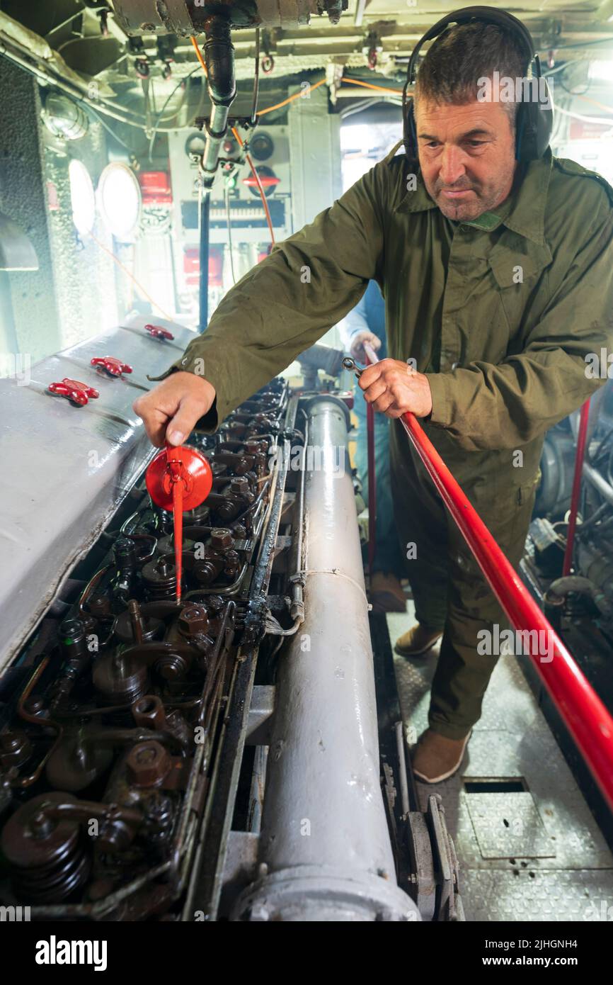 Man, engineer, oiling a large diesel engine in the engine room of the US navy P22 river patrol boat. Cover of engine has been lifted to show pistons. Stock Photo