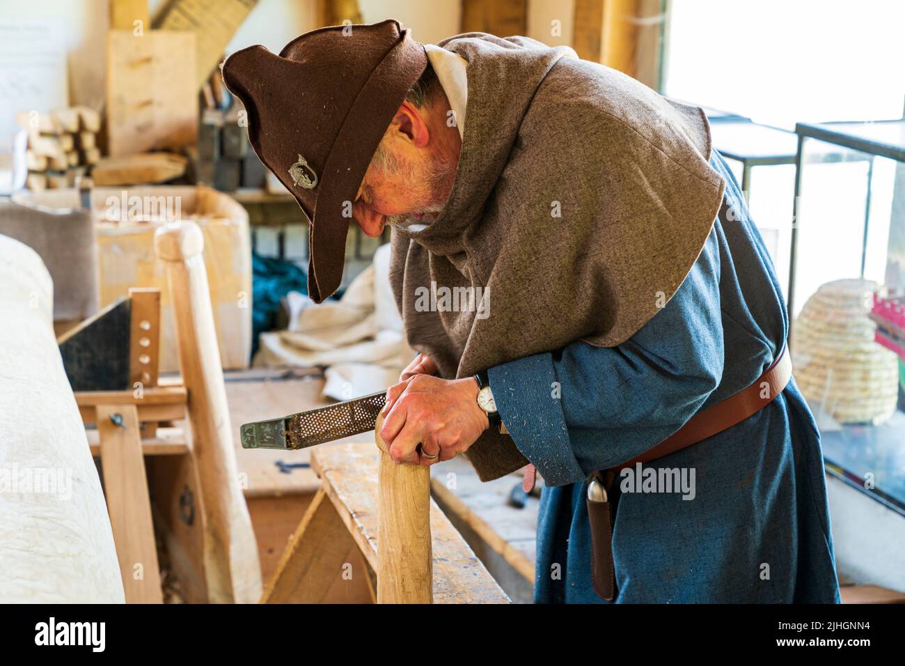 Senior man dressed in Medieval costume of a blue tunic, and brown felt hat, bends over a wooden staff while filing it in a medieval workroom. Stock Photo