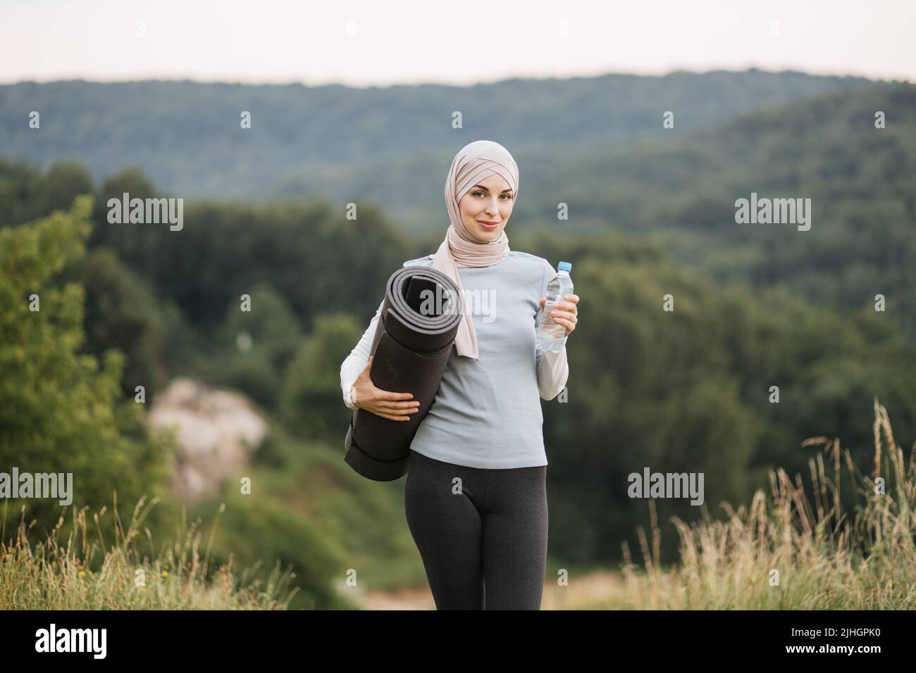 Side view of pretty young woman in hijab and activewear drinking water ...