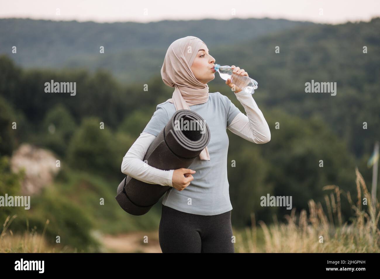 Side view of pretty young woman in hijab and activewear drinking water ...