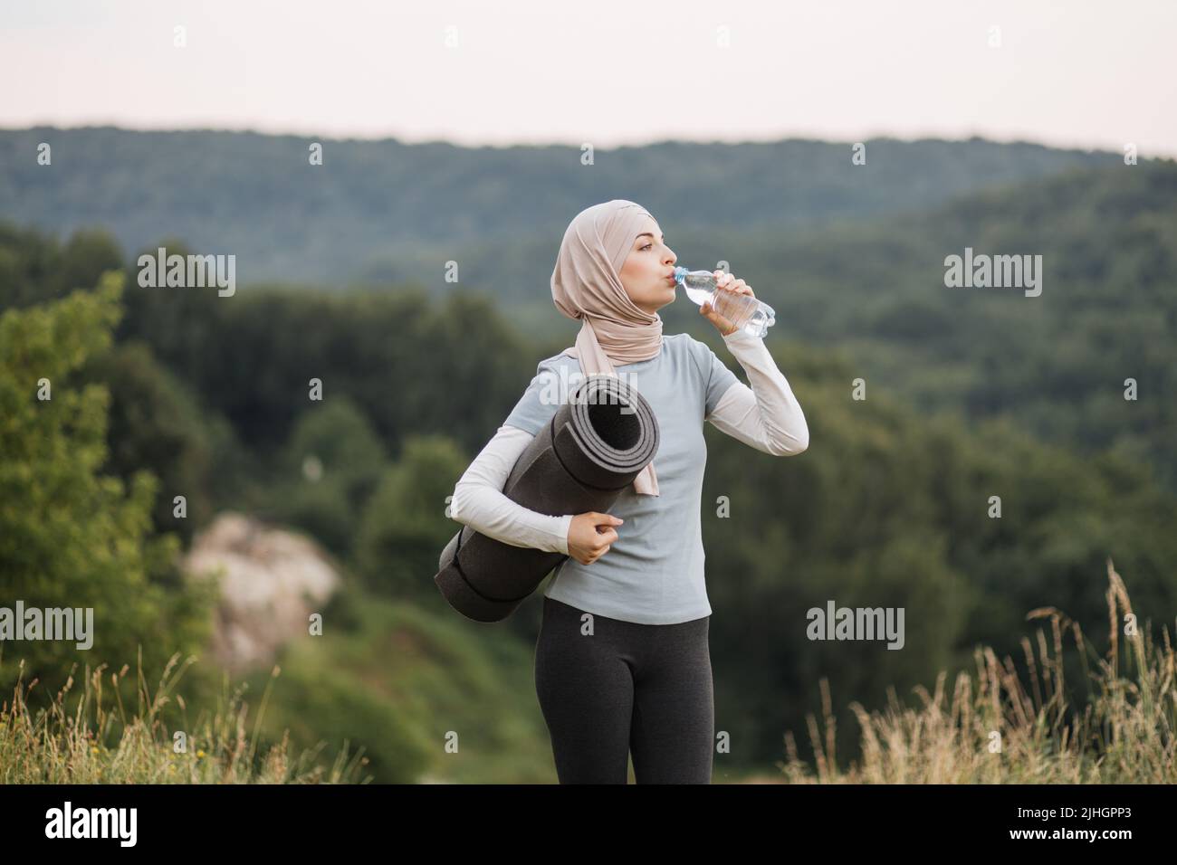 Side view of pretty young woman in hijab and activewear drinking water ...