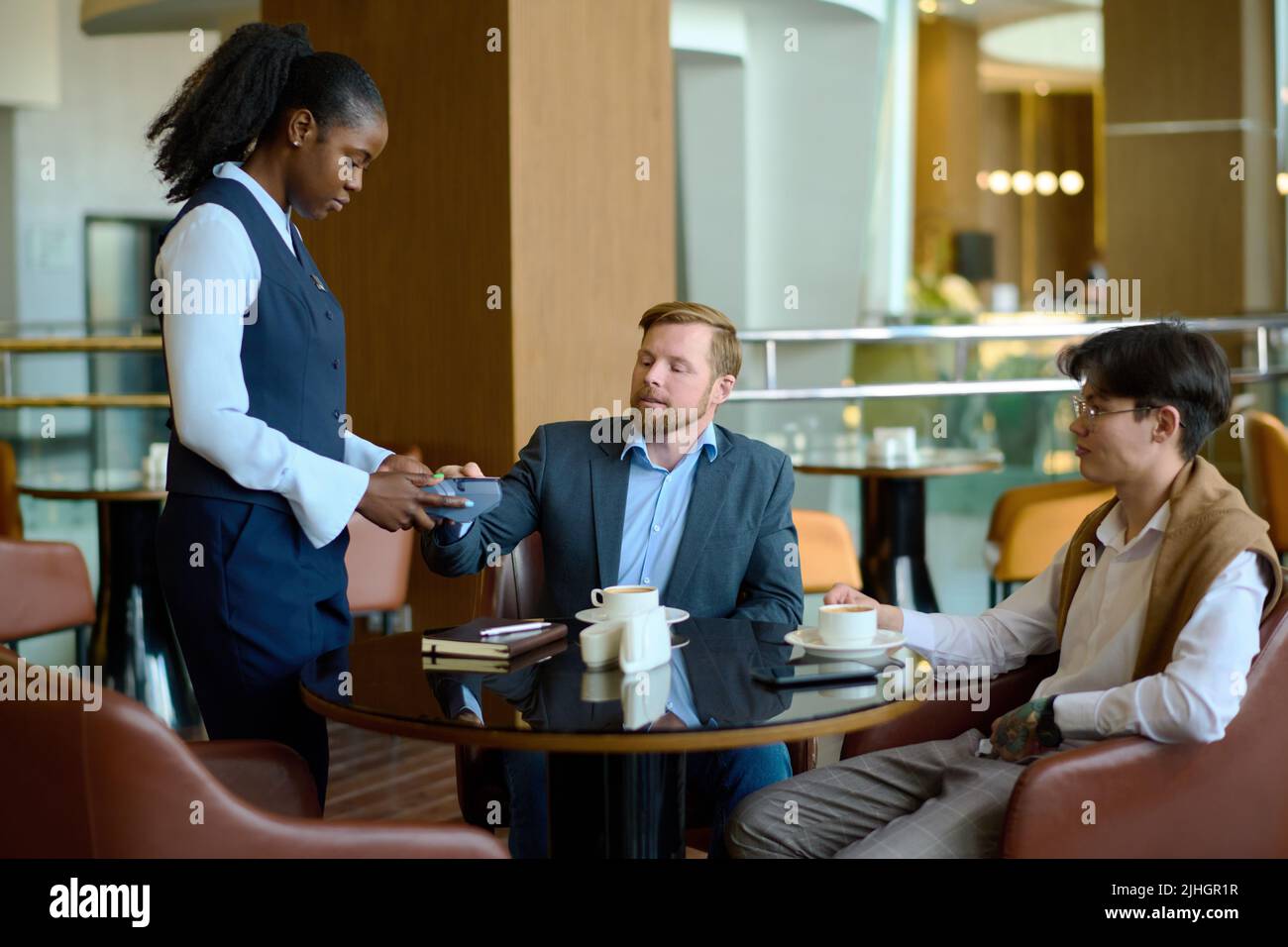 Young elegant businessman paying by credit card for two coffees in cafe while resting by table in the lounge of modern hotel after arrival Stock Photo
