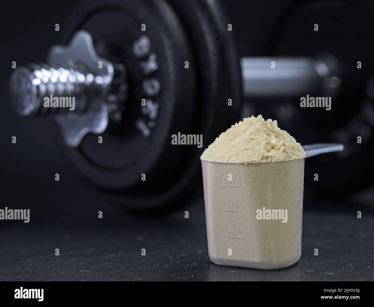 Measuring scoop filled with whey protein powder with dumbbell on black background, protein shake muscle building Stock Photo