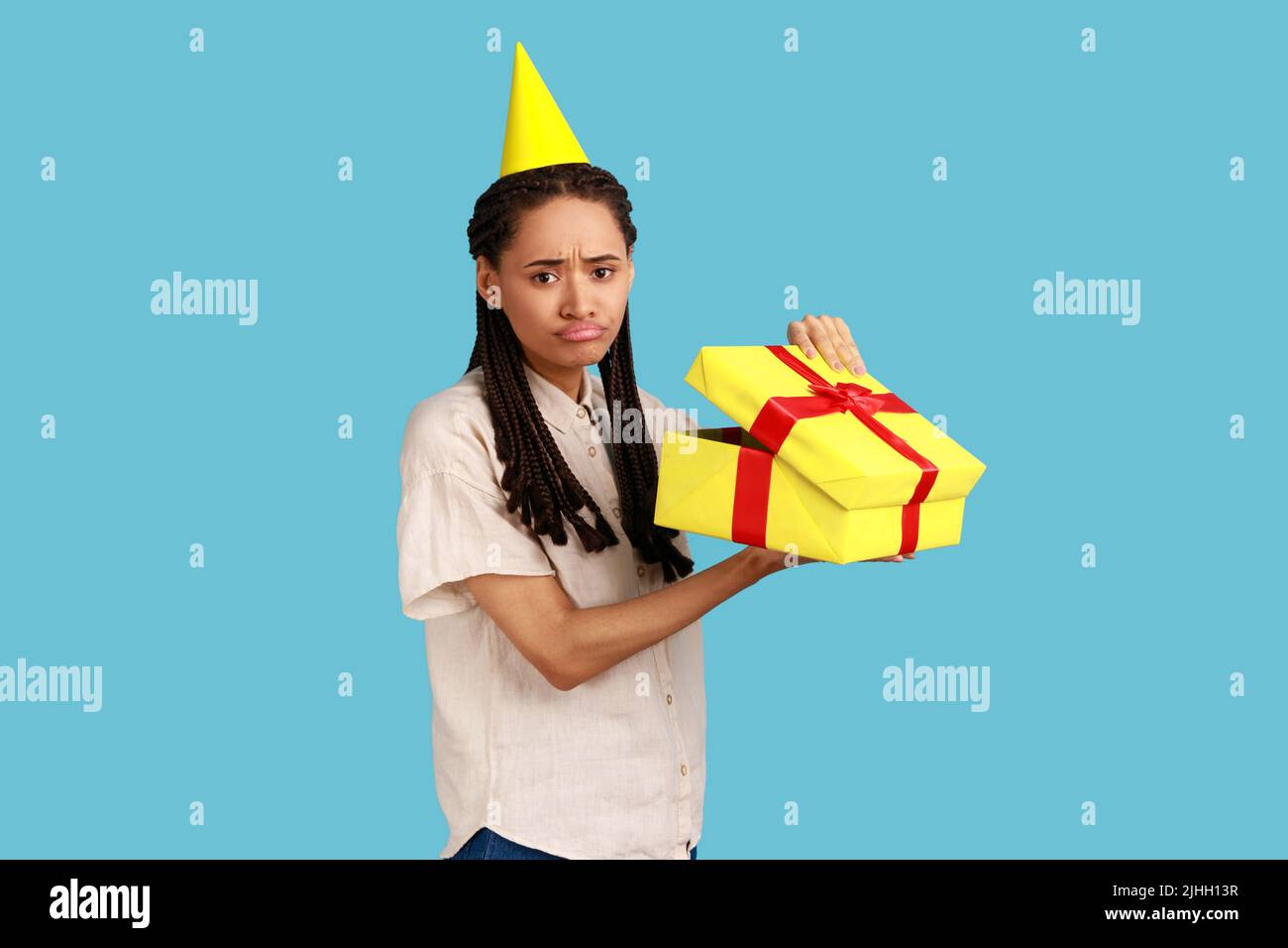 Portrait of unhappy upset woman in yellow party cone looking into gift box, opening present and looking at camera with sadness, wearing white shirt. Indoor studio shot isolated on blue background. Stock Photo