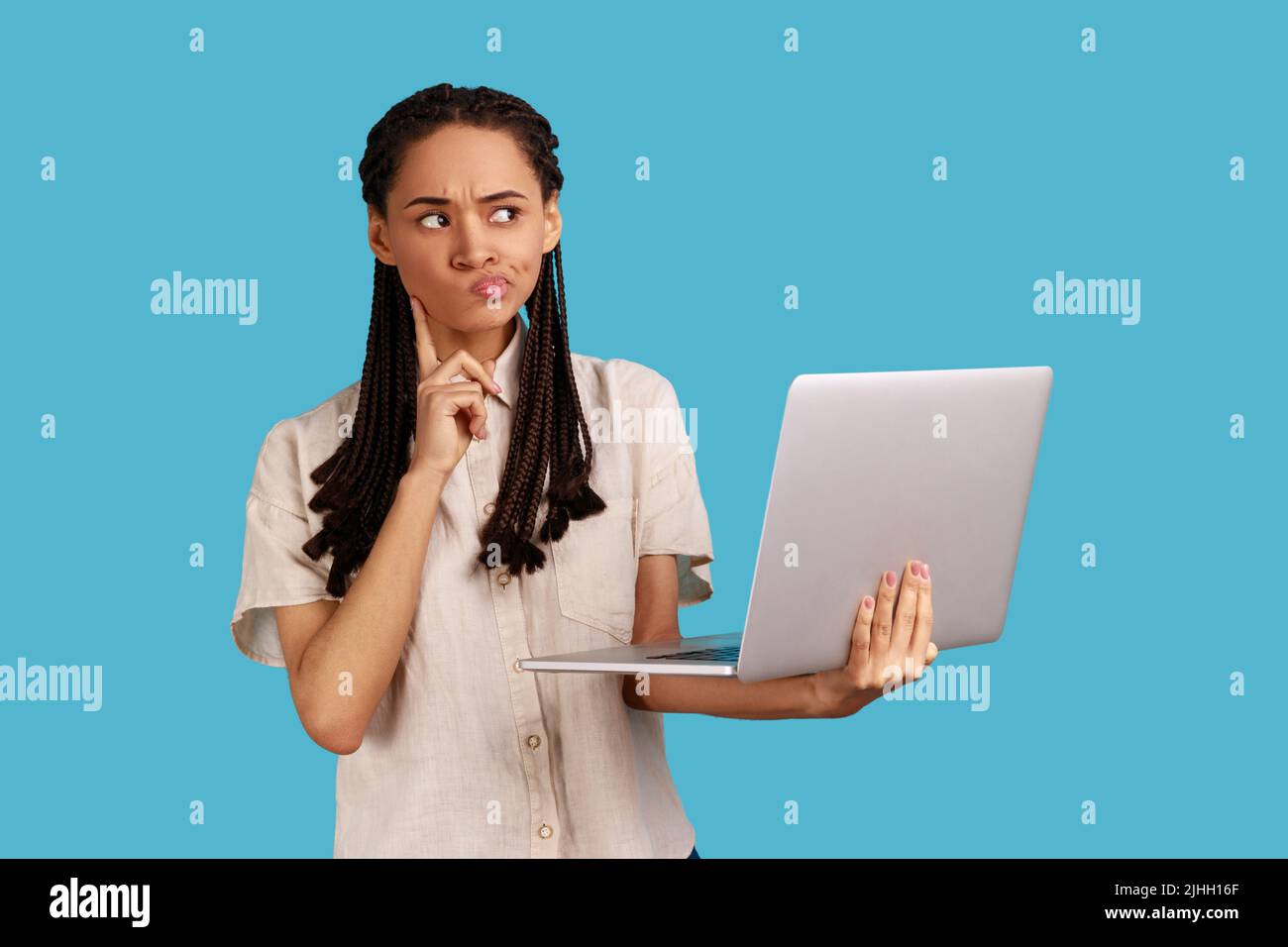 Pensive woman with dreadlocks works on new startup project, makes internet researches, analyzes data, uses modern laptop computer and wireless internet. Indoor studio shot isolated on blue background. Stock Photo