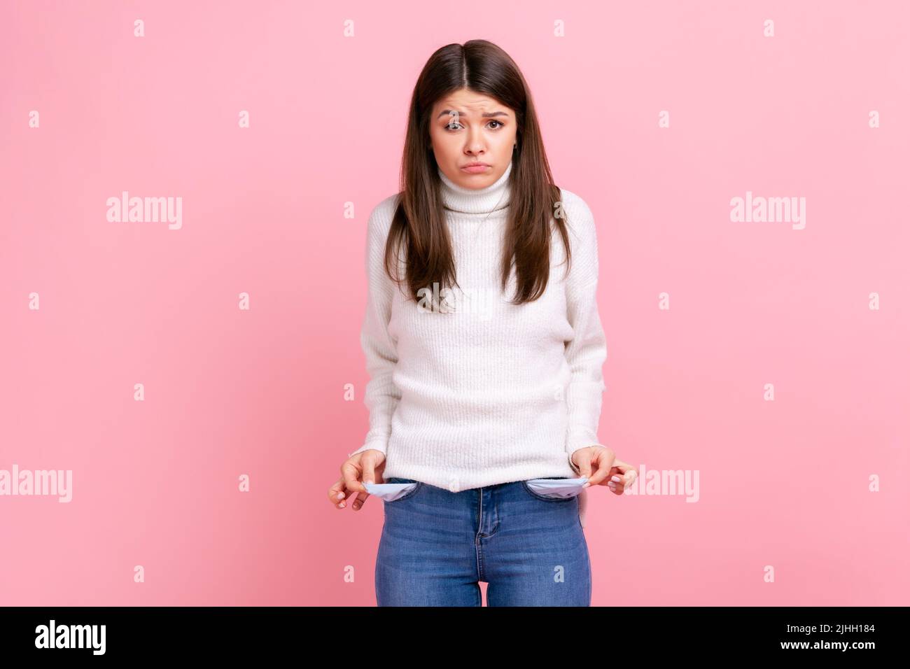 Upset poor girl showing empty pockets and looking frustrated about loans and debts, has no money, wearing white casual style sweater. Indoor studio shot isolated on pink background. Stock Photo