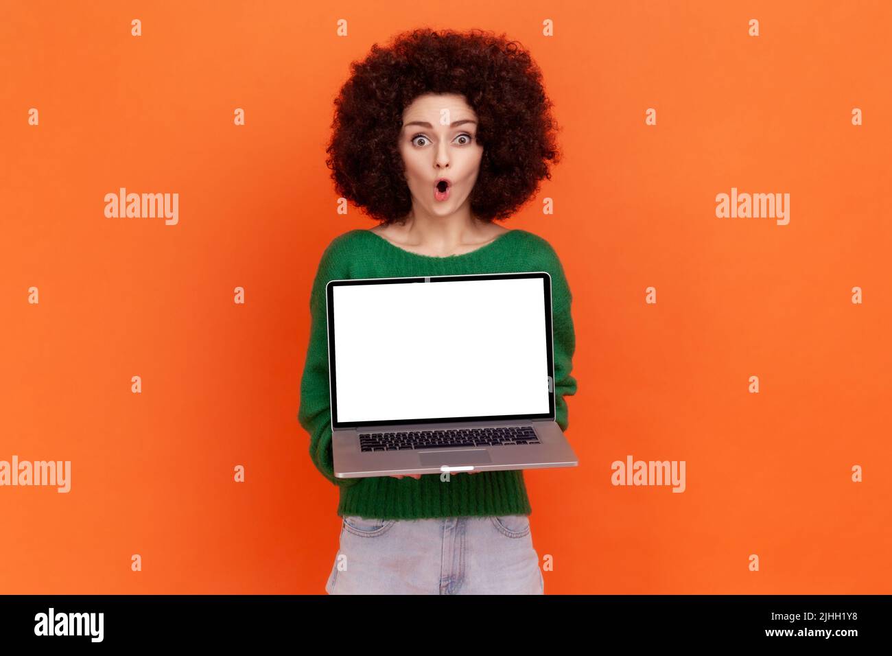 Astonished woman with Afro hairstyle wearing green casual style sweater holding laptop computer with empty screen, showing shocking advertisement. Indoor studio shot isolated on orange background. Stock Photo