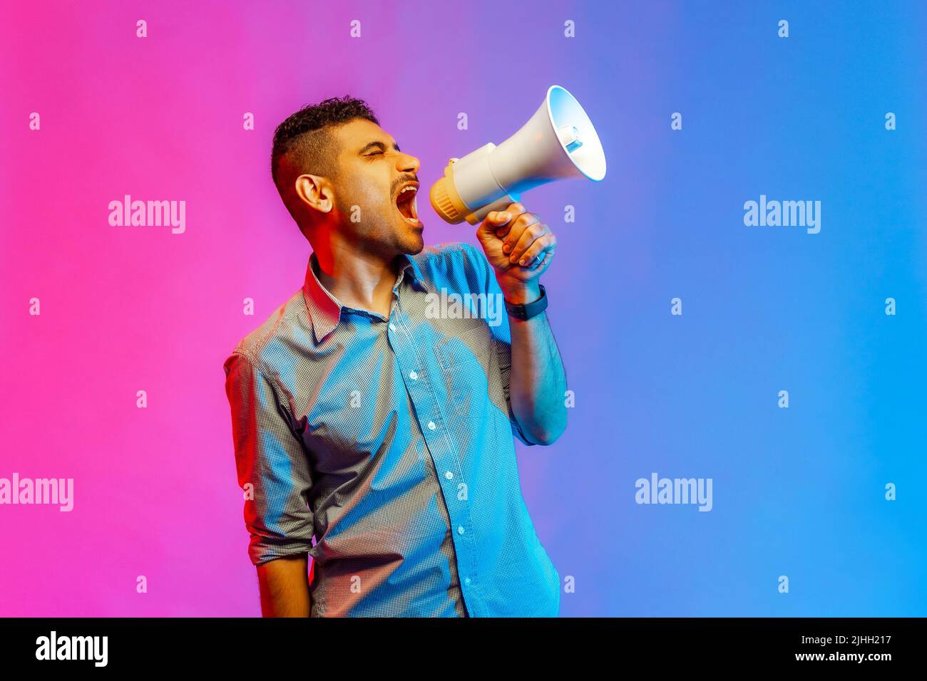 Portrait of aggressive man in shirt screaming at megaphone, making announce, protesting, looking away with angry expression. Indoor studio shot isolated on colorful neon light background. Stock Photo