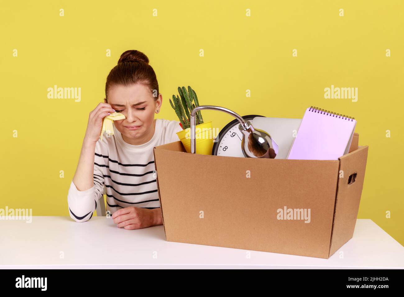 Depressed desperate young woman office worker sitting at workplace with cardboard box with her things and crying, being fired. Indoor studio studio shot isolated on yellow background. Stock Photo