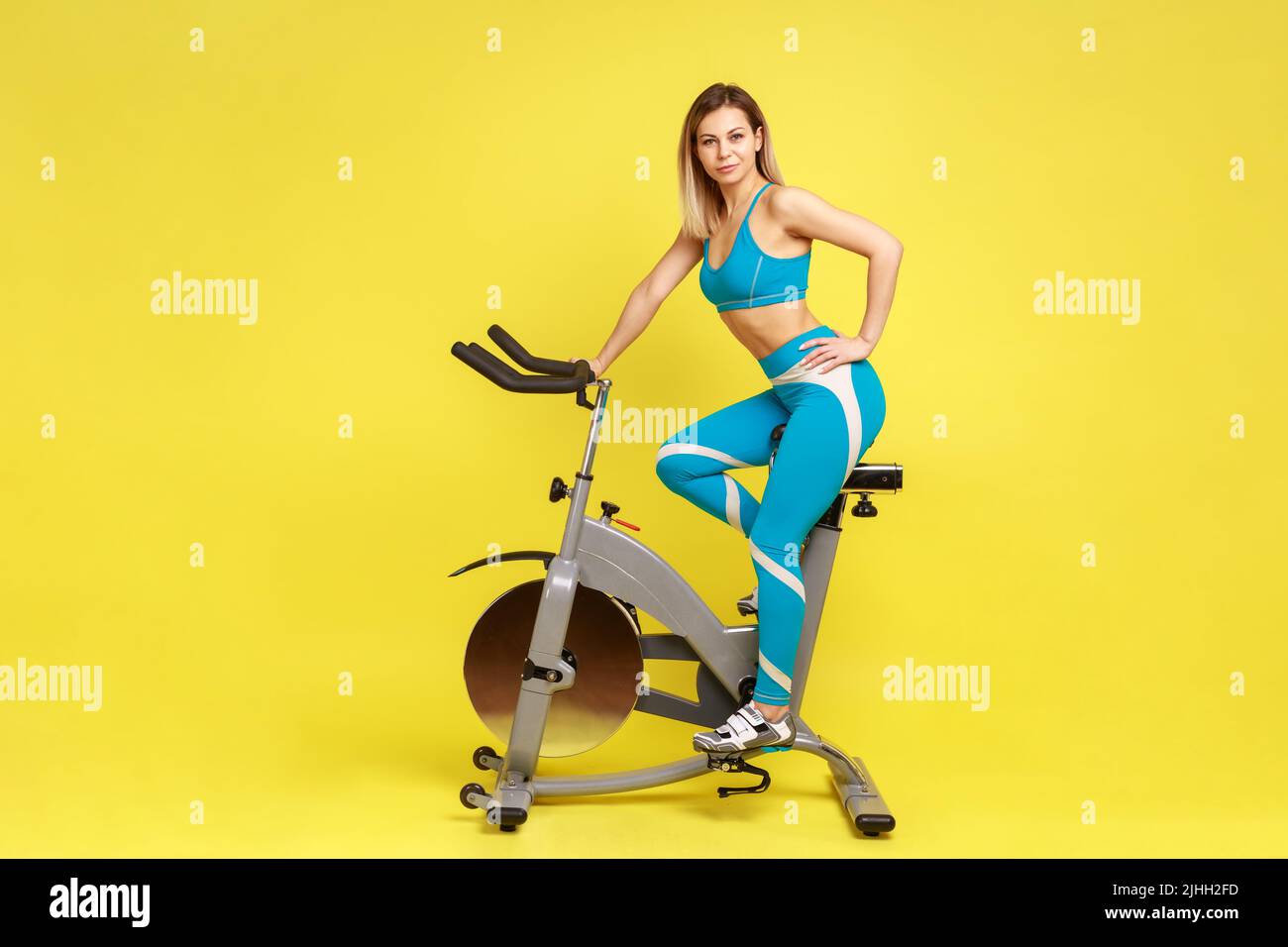 Full length portrait of confident blonde sporty woman on exercise bike, keeping hand on hip, looking at camera, wearing blue sportswear. Indoor studio shot isolated on yellow background. Stock Photo