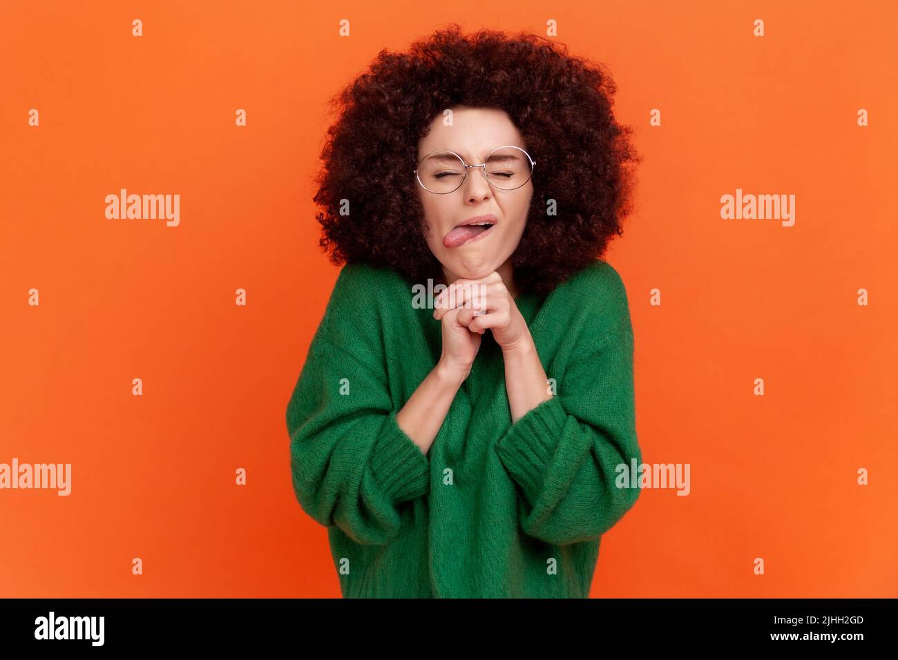 Portrait of funny woman with Afro hairstyle wearing green casual style sweater standing with fists under chin, keeps eyes closed, showing tongue out. Indoor studio shot isolated on orange background. Stock Photo