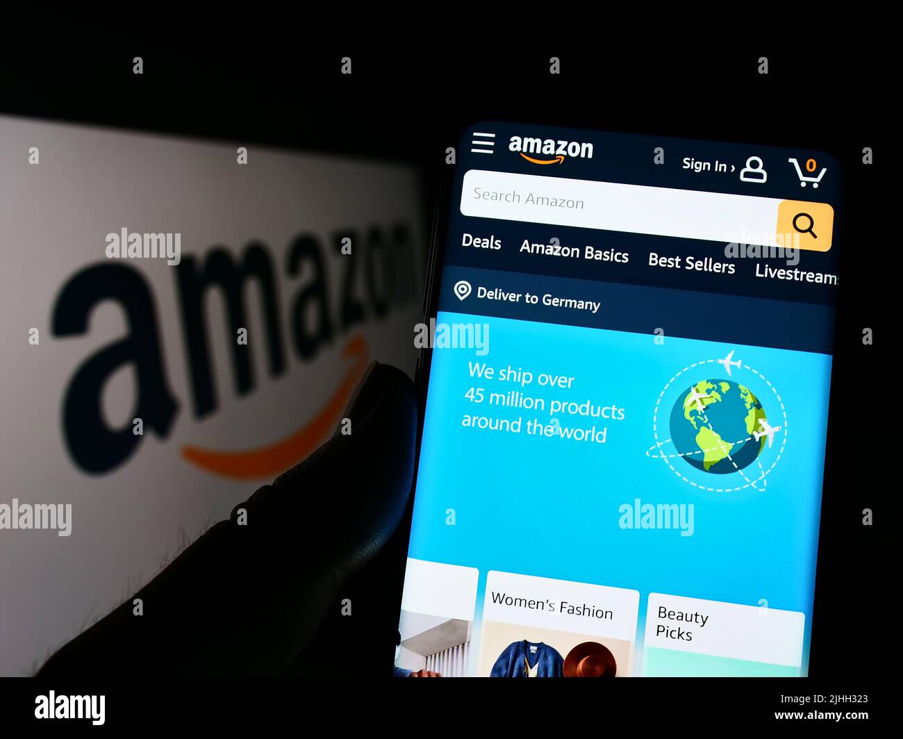 Person holding smartphone with webpage of US technology company Amazon.com Inc. on screen in front of logo. Focus on center of phone display. Stock Photo