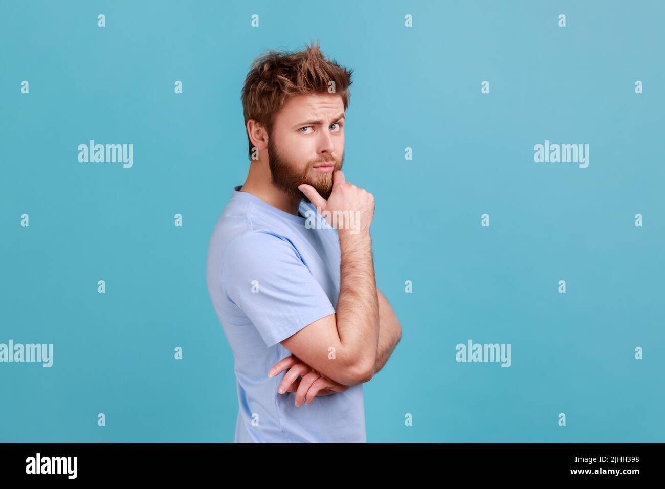 Side view portrait of thoughtful young bearded man touching chin while pondering plan, having doubts about difficult choice, not sure. Indoor studio shot isolated on blue background. Stock Photo