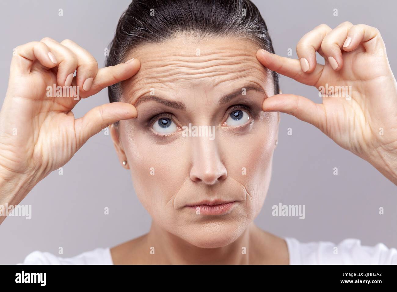 Closeup portrait of middle aged woman checking wrinkles, doing anti aging face yoga exercises to firm and tighten skin and relax muscles on her forehead. Indoor studio shot isolated on gray background Stock Photo