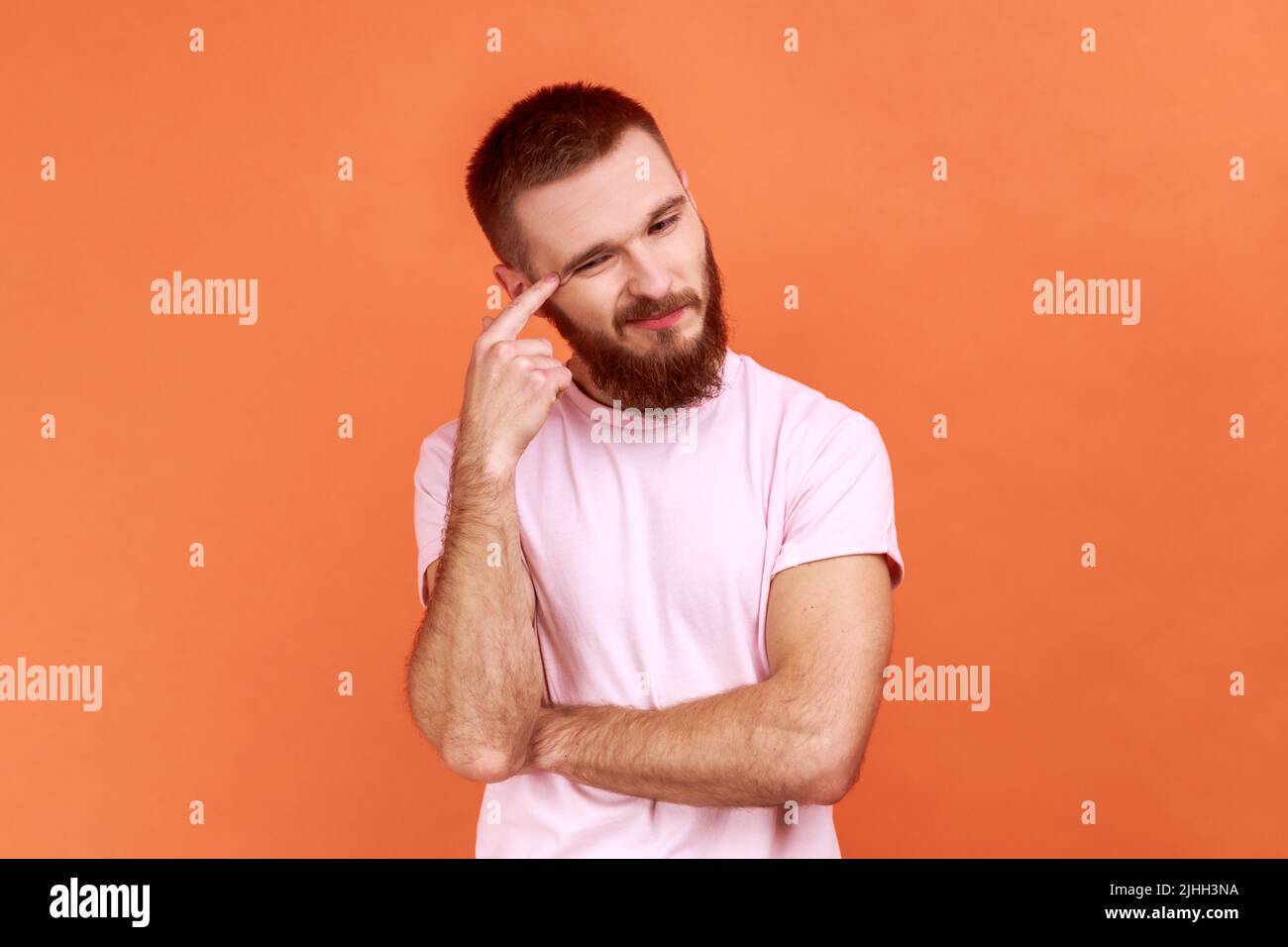 Portrait of bearded man contemplating leaning head on hand, pondering difficult question, trying to make right choice, wearing pink T-shirt. Indoor studio shot isolated on orange background. Stock Photo