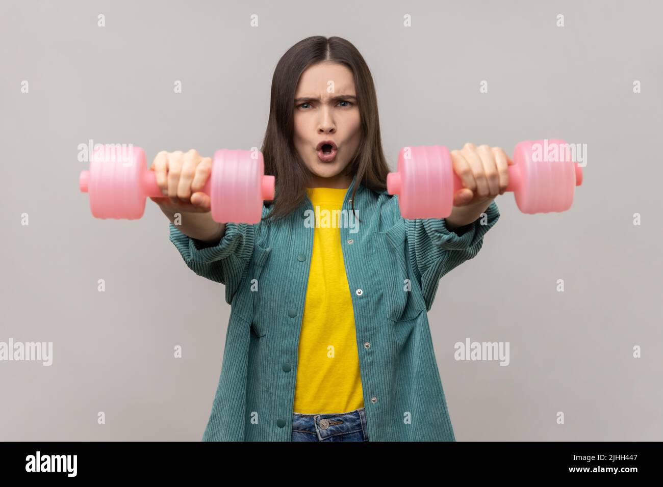 Portrait of woman raising pink dumbbells with effort and looking at camera, pumping up muscles, healthy lifestyle, wearing casual style jacket. Indoor studio shot isolated on gray background. Stock Photo