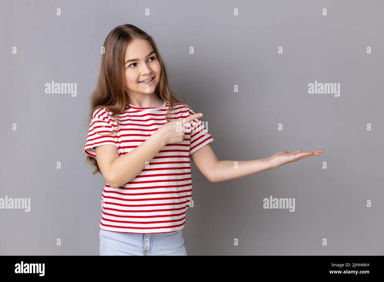 Portrait of little girl wearing striped T-shirt presenting advertising area on her palm and pointing to copy space, holding empty place for commercial. Indoor studio shot isolated on gray background. Stock Photo