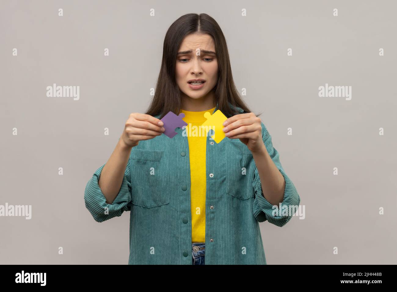 Portrait of scared woman holding colorful pieces of puzzle, two parts of one, symbol of connection and union, association, wearing casual style jacket. Indoor studio shot isolated on gray background. Stock Photo