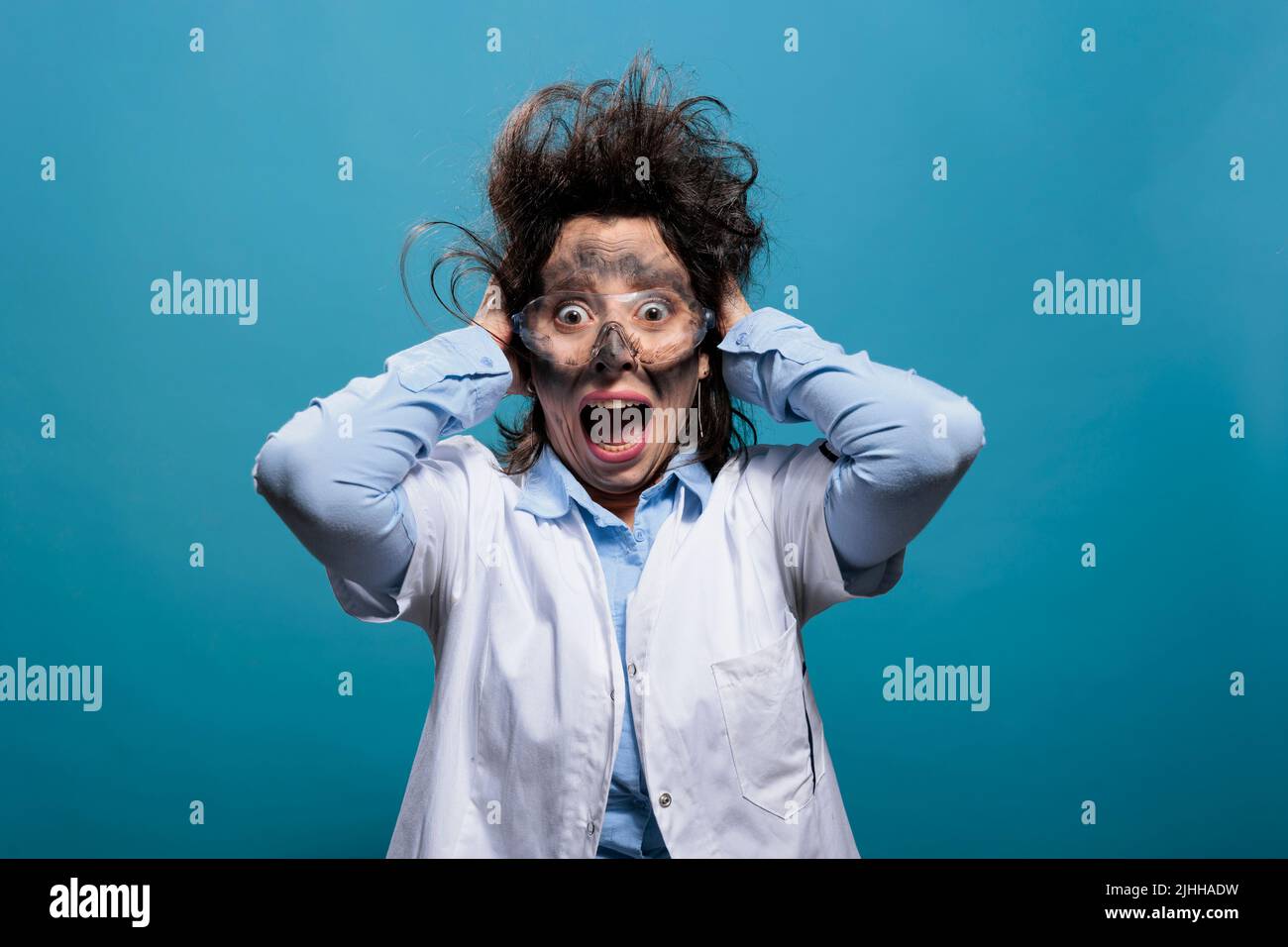 Psycho and scared mad chemist screaming in panic after dangerous chemical explosion and failed scientific experiment. Weirdo scientist with messy hair and dirty face shouting on blue background. Stock Photo