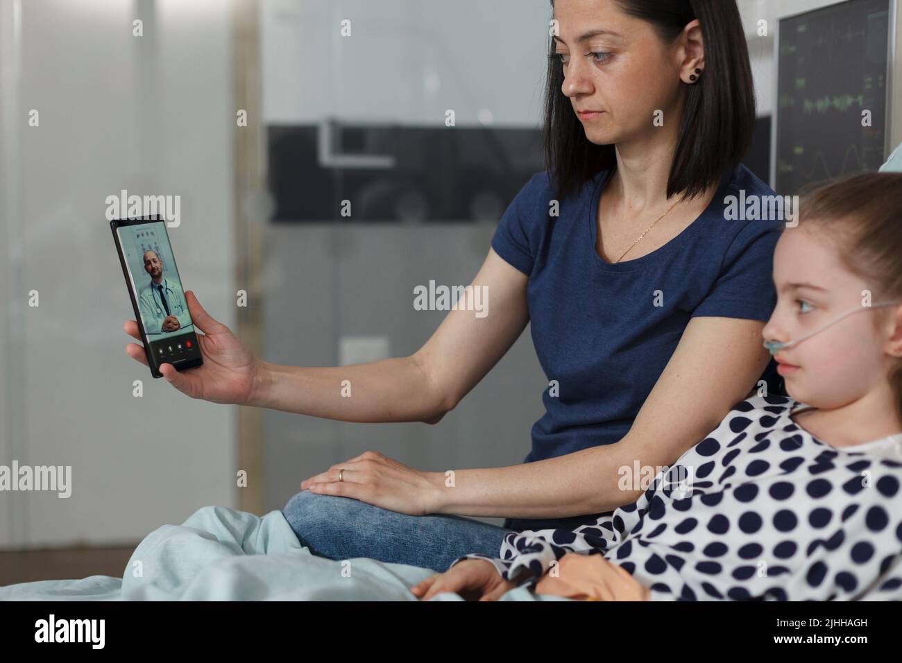 Ill kid and parent discussing with physician on smartphone virtual online call. Mother and sick daughter talking with doctor on telehealth phone videocall while sitting in hospital pediatrics ward. Stock Photo