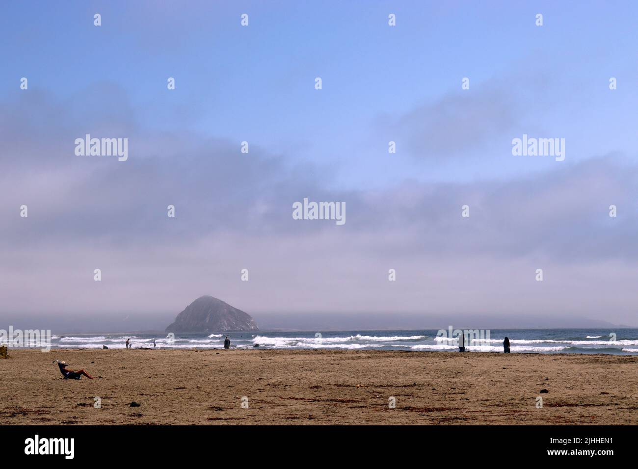 A calm, slightly overcast afternoon on a beach near Cayucos, CA with few people and gentle waves. Morro Bay sits in the distance with its giant rock. Stock Photo