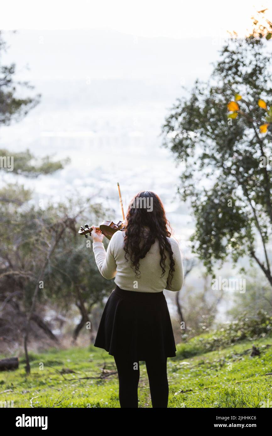 Brunette woman seen from back playing violin outside in the mountain. Vertical Stock Photo