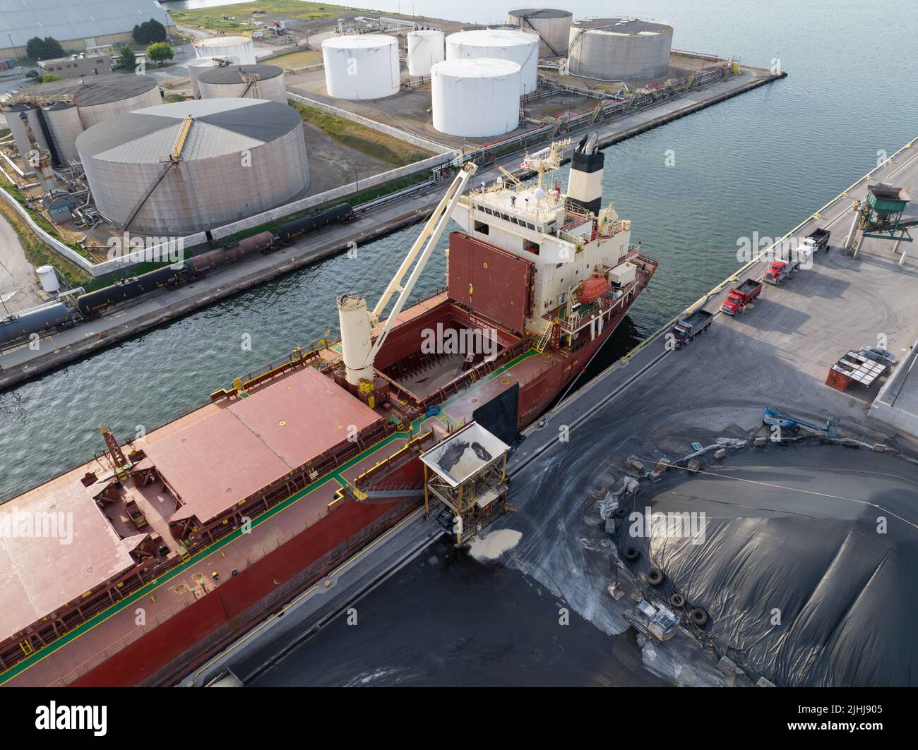 A bulk carrier cargo ship is seen at port offloading dry goods by crane, filling numerous dump trucks during the day. Stock Photo