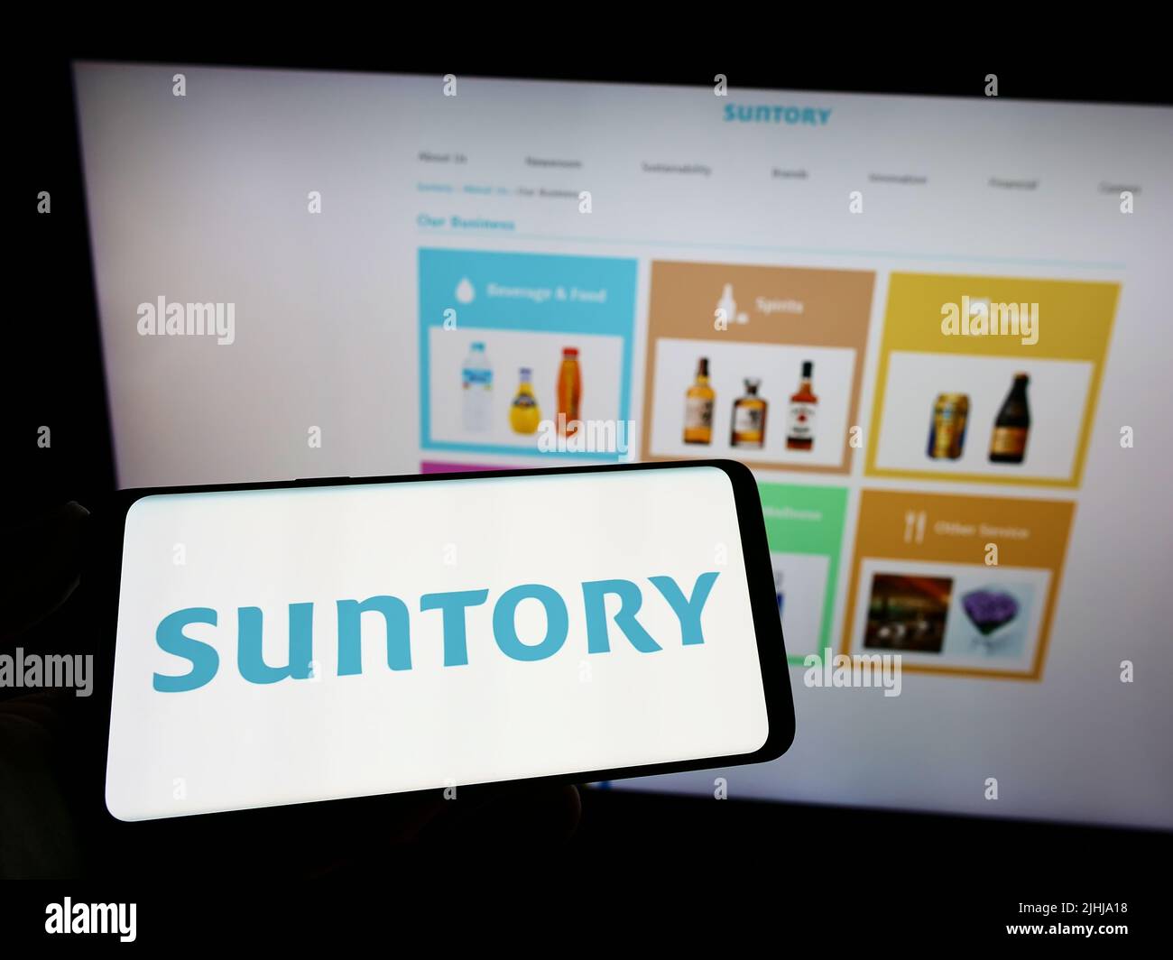 Person holding mobile phone with logo of Japanese beverage company Suntory Holdings KK on screen in front of web page. Focus on phone display. Stock Photo