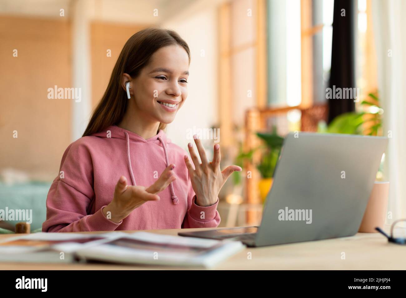 Cheerful schoolgirl in earphones having video call on laptop, sitting at table in bedroom and gesturing at webcam Stock Photo