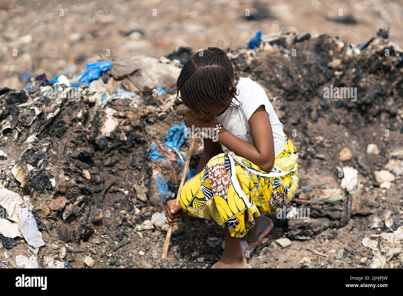 Tiny African girl rummaging through a pile of damp and dirty street litter with a wooden stick, looking for recyclable items to sell Stock Photo