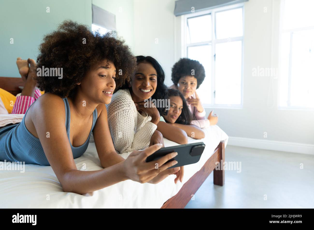 Biracial woman with afro hair taking selfie over cellphone with friends lying side by side on bed Stock Photo