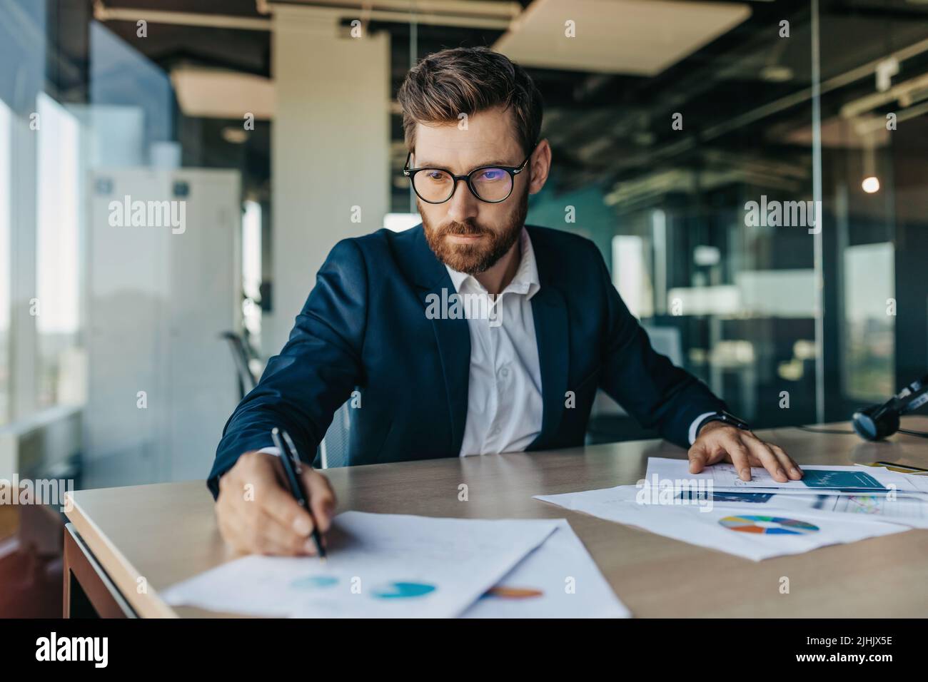 Concentrated handsome businessman in glasses working with documents in office interior, checking and signing papers Stock Photo