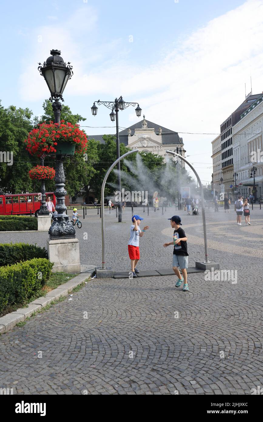 Two little boys enjoy refreshment from public nebulizers on a city square; water mist cooling, fog cooling, water sprayers Stock Photo
