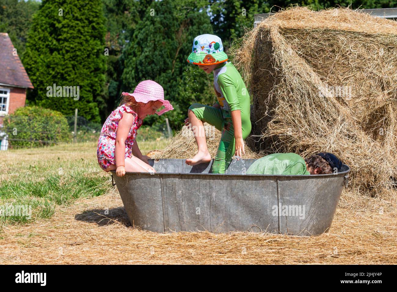 Kidderminster, Worcestershire, UK. 19th July, 2022. During a break from optional home schooling due to the heatwave, Henley Mills, 9, with brother Reggie, 6, and sister Myla-May, 3, have a splashing time in an old tin bath trying to keep cool in the family's farmyard near Kidderminster, Worcestershire, in a morning where temperatures are set to become UK record highs. Credit: Peter Lopeman/Alamy Live News Stock Photo