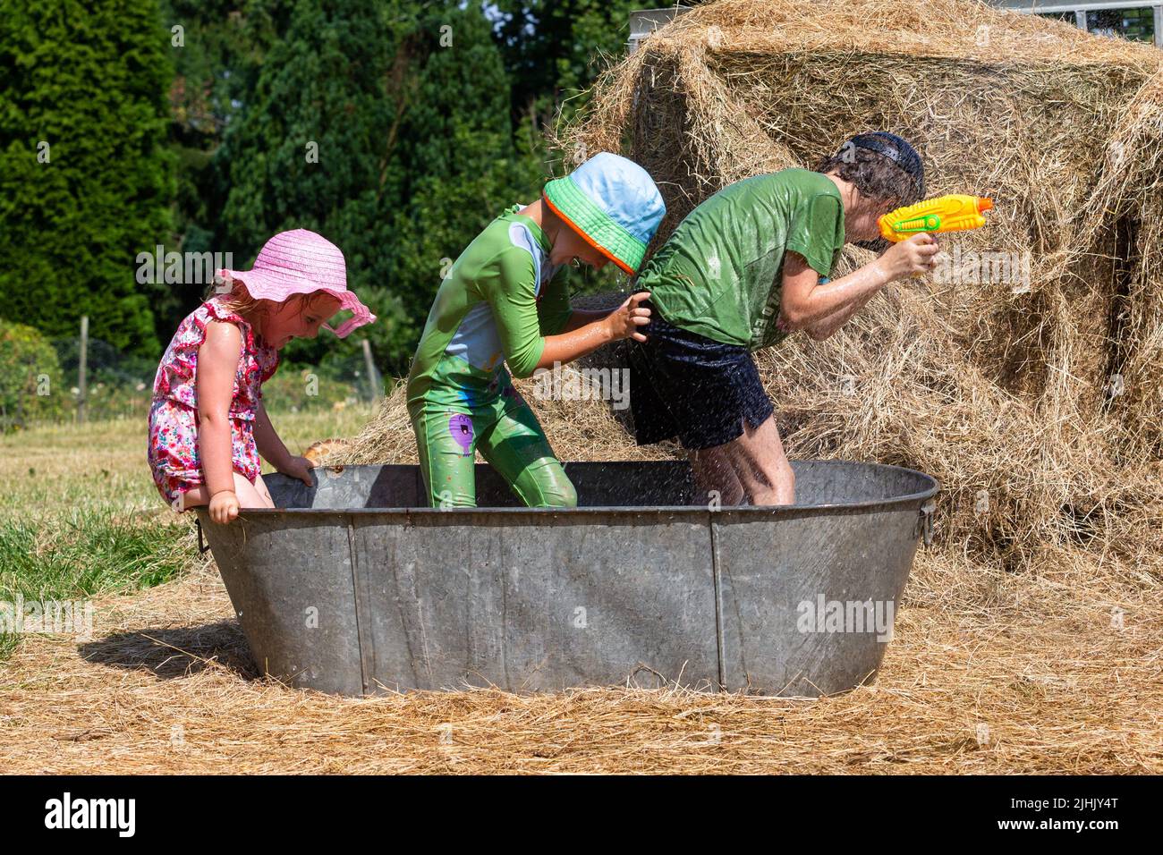 Kidderminster, Worcestershire, UK. 19th July, 2022. During a break from optional home schooling due to the heatwave, Henley Mills, 9, with brother Reggie, 6, and sister Myla-May, 3, have a splashing time in an old tin bath trying to keep cool in the family's farmyard near Kidderminster, Worcestershire, in a morning where temperatures are set to become UK record highs. Credit: Peter Lopeman/Alamy Live News Stock Photo