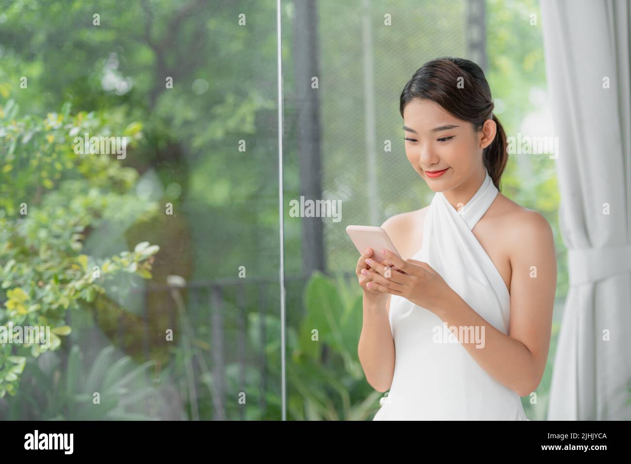 Asian girl sits at the table next the window holding a mobile phone and looking at the screen Stock Photo