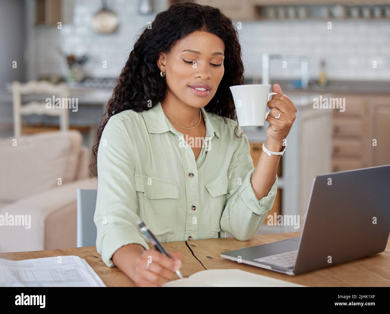 Working from home is so comfortable for me. a young woman writing notes and drinking coffee while working on a laptop at home. Stock Photo