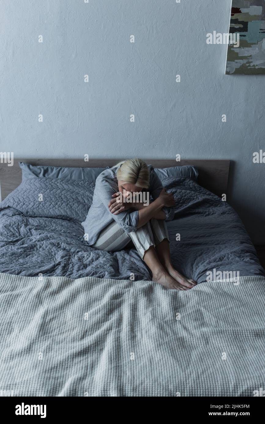 blonde woman hugging knees on bed while feeling pain in stomach during