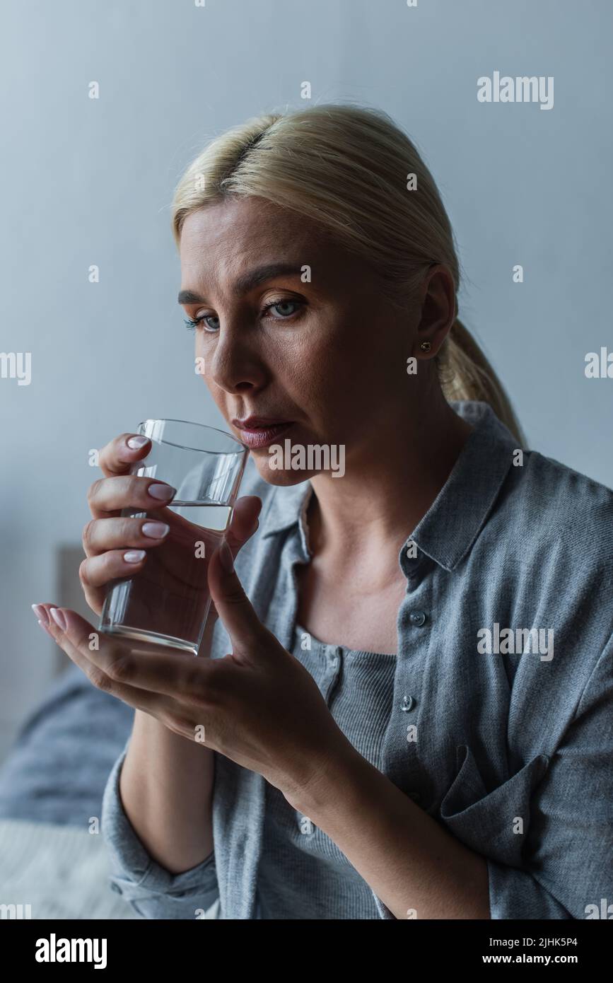 sad blonde woman with menopause drinking fresh water from glass Stock Photo